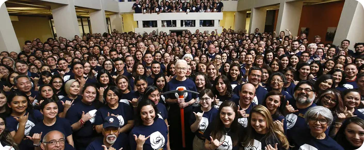 Diana Natalicio, in the middle of the hall, holding a pick-axe, surrounded by students and staff in UT El Paso colors and wear.