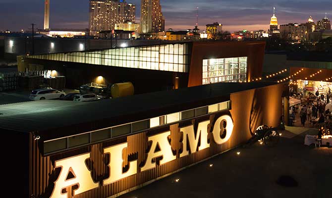 Nightlife photo of the city of San Antonio with the facade of the word Alamo lit up in lights.
