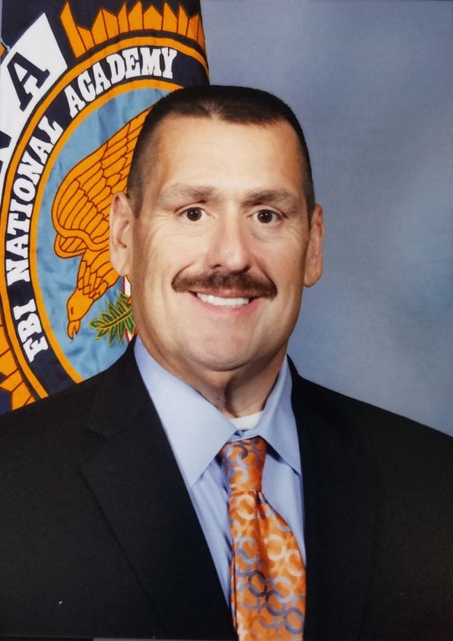 Charlie Patnode smiles in front of a flag with a seal that reads, in part, 'FBI National Academy'.