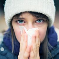 close-up photo of a woman in warm clothes and sneezing into a Kleenex.